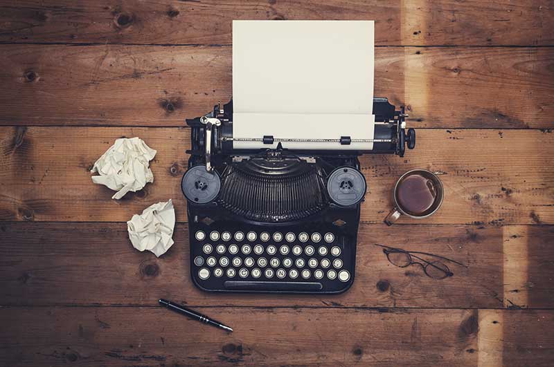 A typewriter on a wooden table with crumpled pieces of paper, a pen, a mug of coffee and a pair of glasses beside it