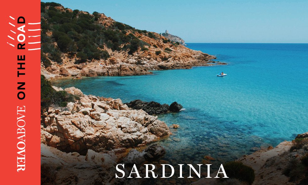 Bright blue water and a rocky coastline in Sardinia, Italy, with the words “Overabove on the Road” on the side